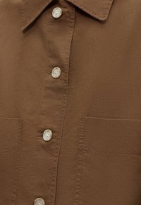 Καφέ υφασμάτινο πουκάμισο με γιακά, λευκά κουμπιά, ορατές ραφές και τσέπη στη δεξιά πλευρά της πατιλέτας.