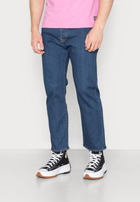 Fatos de ganga de denim azul escuro com um corte reto, usados com ténis de cano alto pretos que apresentam accentos brancos e solas de borracha de goma.