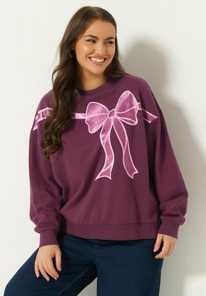 Lächelnde Frau, die ein lila Sweatshirt mit großem pinkglitzerndem Schleifenmotiv trägt, steht mit einer Hand auf ihrem Oberschenkel gegen eine weiße Wand.