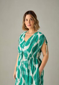 Robe à motifs verts et blancs avec un décolleté en V, des manches courtes, une taille à cordon et des poches latérales. Comprend des liens aux extrémités des manches.