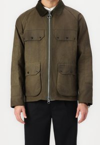 Mann mit brauner Zip-Jacke, die über vier knöpffällige Taschen und einen Cordkragen verfügt, kombiniert mit schwarzen Hosen.