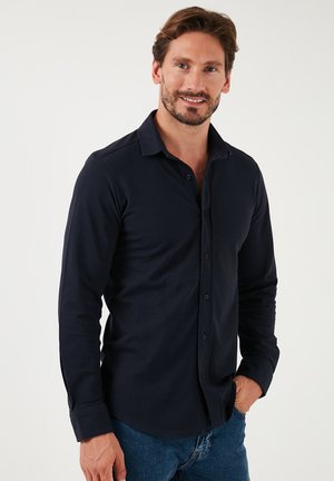Camisa de manga larga azul marino hecha de un material suave, con cuello abotonado, botones en la parte frontal y un dobladillo curvado. Combinada con vaqueros.