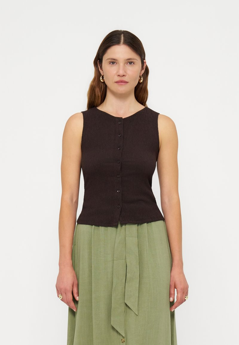 Femme de face portant un haut sans manches marron foncé boutonné et une jupe vert clair taille haute avec un détail nœud à l'avant.