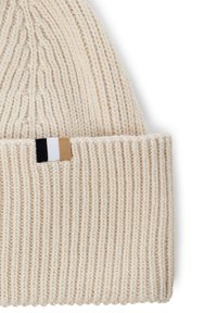 Knitted cream beanie with a folded ribbed cuff. Features a small tag with black, white, and beige stripes. Soft texture and rounded shape.