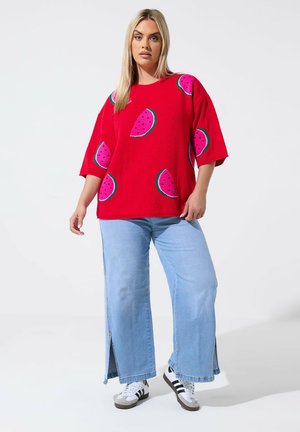 Femme portant un pull rouge avec des tranches de pastèque, un jean large bleu clair avec des fentes sur les côtés et des baskets blanches avec des bandes noires.