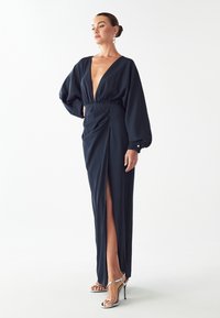 Námornícky modré maxi šaty s hlbokým V výstrihom, balónovými rukávmi a bočným rozparkem. Obsahuje naťahované detaily v oblasti pásu a hladký povrch látky.