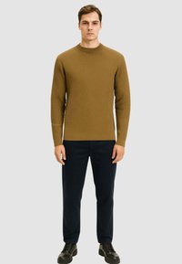 Maglione a coste color khaki con scollo rotondo, abbinato a pantaloni navy e scarpe nere. Il modello si trova su uno sfondo grigio neutro.