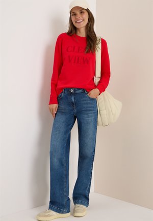 Sweat-shirt rouge avec l'inscription "CLEAN VIEW", jeans bleu taille haute à jambes larges, baskets beiges et sac à épaule crème sur fond neutre.