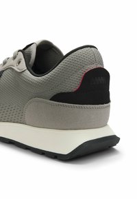 Gros plan sur le talon d'une basket grise en textile et daim, avec des accents noirs et rouges et une semelle en caoutchouc blanche.