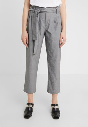 Kobieta w szarych, krótkich spodniach z wysokim stanem i materiałowym paskiem wiązanym w kokardę oraz czarnych mokasynach na kwadratowym nosku z srebrnym łańcuszkiem.