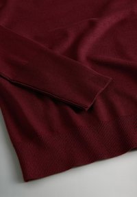 Burgundi värvi kootud sall, millel on ribakujulised varrukad ja ääred, sile tekstuur ja keha külge püüdlik disain. Kude näib olevat kerge ja pehme.