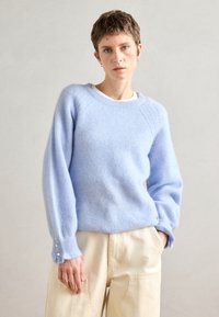Lyseblå, fluffy strikkegenser med senkede skuldre, ribbet tekstur og knappdetaljer på ermene, kombinert med beige bukser.