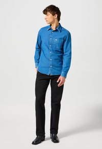 Blå denimskjorta med knäppning, långa ärmar, vänster bröstficka och svarta byxor. Modellen bär svarta klackstövlar och står lite vänd åt vänster.