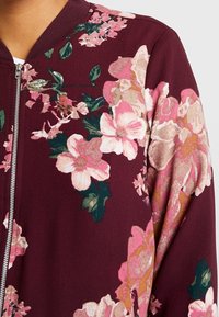 Bordowy bomber z gładkiego materiału, zdobiony kwiatowym wzorem w odcieniach różu i zieleni, z zamkiem błyskawicznym w kolorze srebra.