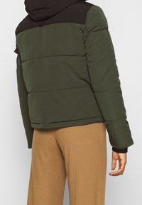 Persona che indossa un giubbotto di piumino verde bicolore con cappuccio e pantaloni a coste beige, fotografata da dietro.