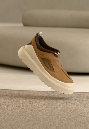 Chaussure à enfiler en daim marron et mesh avec une semelle épaisse crème, présentant un design élégant et minimaliste avec des accents métalliques.