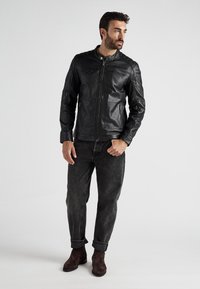 Mann in einer schwarzen Lederjacke, dunkelgrauen Jeans mit hochgerollten Ärmeln und dunkelbraunen Wildlederstiefeln, der vor einem einfarbigen hellen Hintergrund steht.