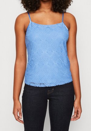 Frau trägt ein hellblaues Spitzen-ärmelloses Oberteil mit dünnen Trägern und schwarze eng anliegende Jeans vor einem schlichten hellen Hintergrund.