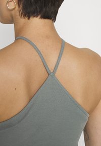 Top de alças cinza com nervuras e finas alças cruzadas nas costas; textura suave e design ajustado, mostrando o tom de pele e tatuagens no ombro.