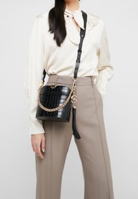 Borsa a secchio nera in pelle goffrata a forma di coccodrillo con dettagli in catena dorata e un pon-pon. Presenta una tracolla removibile e una texture in pelle liscia.
