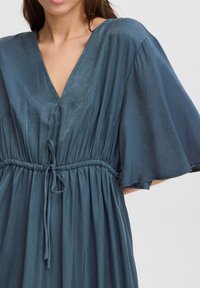 Fließendes aquafarbenes Kleid aus leichtem Stoff; mit tiefem V-Ausschnitt, kurzen, flatternden Ärmeln, elastischem Bund und einer Bindetechnik.
