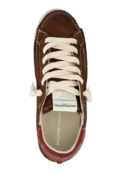 Sneaker in suede marrone con lacci bianchi, suola in gomma e accento rosso vicino al tallone. Presenta dettagli cuciti e un'etichetta in tessuto all'interno.