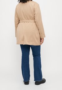 Beżowy trencz z paskiem, o długości midi, gładkiej fakturze i długich rękawach. Noszony na niebieskich dzwonach i czarnych botkach sięgających kostek.