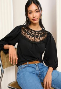 Černý top s krajkovou vyšívanou předsádkou a balónovými rukávy, kombinovaný s modrými džíny. Modelka sedí, má hnědý opasek a uvolněný postoj.