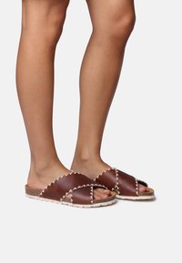 Brown leather slide sandals with cross strap design, scalloped edges, and a textured cork footbed. White stitching accents the edges.