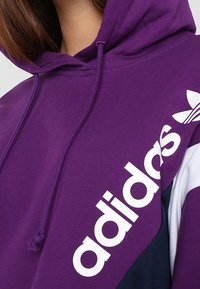 Felpa Adidas viola con logo bianco ben visibile, cappuccio con cordino e design a blocchi di colore con accenti blu navy e bianchi. Tessuto morbido.