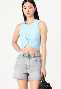 Top corto senza maniche di colore azzurro chiaro con un nodo frontale, abbinato a pantaloni corti in denim grigi a vita alta. I pantaloni hanno un aspetto sbiadito e consunto.