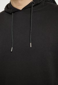 Černá mikina s kapucí a šňůrkou. Hladký povrch látky, splétané černé šňůrky a kovové konce šňůrek.