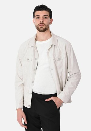 Uomo con capelli scuri e barba, che indossa una giacca beige chiaro, camicia bianca e pantaloni neri, in piedi davanti a uno sfondo bianco.