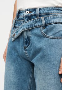 Vaqueros de mezclilla azul claro con una textura ligeramente desteñida, que presentan un cierre de botones, trabillas para cinturón y costuras visibles a lo largo de las uniones.