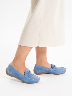 Legs wearing light blue suede loafers with metal bit detail, paired with cream-colored wide leg pants against white background.