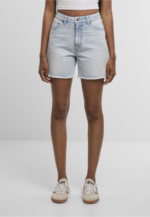 Személy világoskék farmer rövidnadrágot és bézs sportcipőt visel, egyszínű szürke háttér előtt áll.