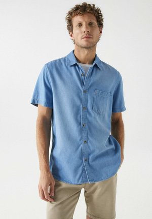 Camisa de denim de manga corta, de color azul claro, con cuello abotonado, un bolsillo en el pecho y costuras tonales. Textura de tela de algodón suave.