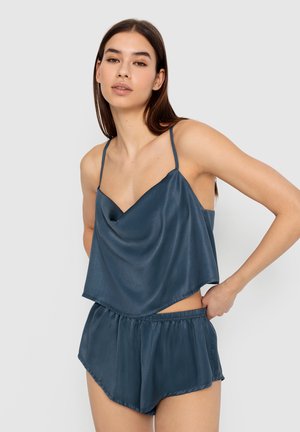 Satinový pyžamový set v hlbokej teal farbe, ktorý obsahuje voľný tank top s ozdobným golierom a tenkými ramienkami, ako aj zodpovedajúce šortky s elastickým pásom a zakriveným lemom.