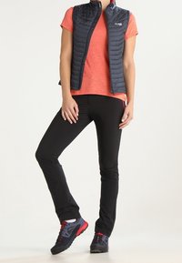 Veste matelassée noire portée sur une chemise orange, associée à un pantalon noir et des chaussures avec des accents rouge foncé. Le matériau est lisse ; la coupe est athlétique.