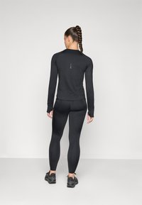 Svart langermet topp og leggings laget av elastisk stoff. Minimalistisk design med en logo på baksiden. Modellen har på seg svarte treningssko.