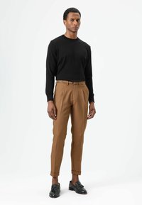 Zwarte trui met lange mouwen, lichtbruine op maat gemaakte broek met zichtbare plooien, zijzakken en een slanke pasvorm. Zwarte loafers maken de look compleet.