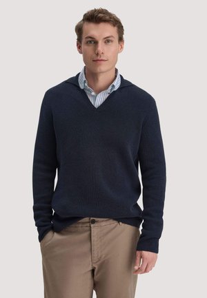 Junger Mann mit hellbraunen Haaren, trägt einen dunkelblauen V-Ausschnitt-Pullover über einem blau gestreiften Hemd und beigefarbenen Hosen, Hände in den Taschen, neutraler Hintergrund.