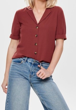 Woman wearing a short-sleeve rust-colored button-up blouse tucked into high-waist light blue jeans, posed with one hand near the waistband.