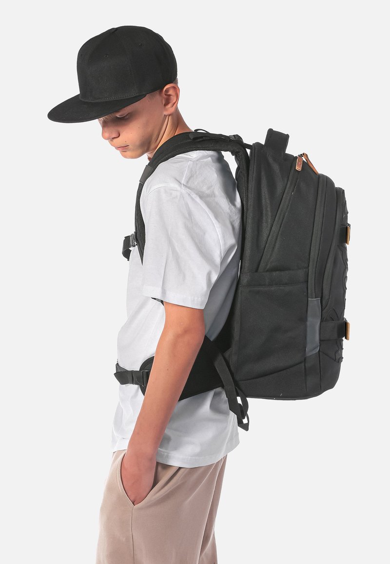 Zaino in tessuto nero con più scomparti e spallacci regolabili, indossato sopra una camicia bianca. Presenta un design robusto e dettagli decorativi.