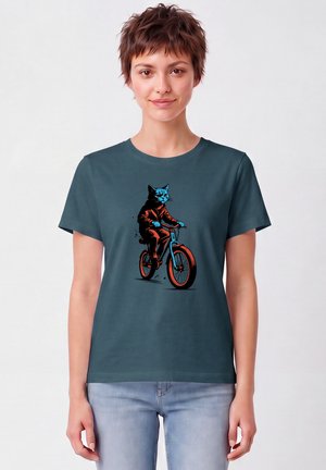 Frau mit kurzen Haaren, die ein dunkel-türkisfarbenes T-Shirt mit einer Grafik einer Katze im Anzug trägt, die ein blau-orangefarbenes Fahrrad fährt.