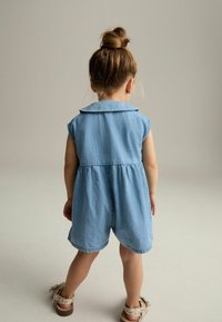 Combinaison en denim bleu clair avec des manches courtes, un col et une taille froncée. Portée avec des sandales à motifs marron et beige.