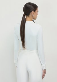 Weißes Langarmshirt mit Kragen und Knopfmanschetten. Kombiniert mit hochgeschnittenen weißen Hosen, was eine elegante Silhouette erzeugt.