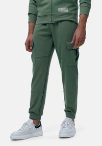 Zelené jogger nohavice vyrobené z mäkkého, elastického materiálu s dvoma bočnými vreckami a elastickými manžetami, skombinované s bielymi teniskami.