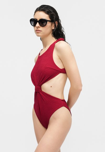 Γυναίκα που φοράει υπερμεγέθη μαύρα γυαλιά ηλίου και μια κόκκινη ολόσωμη μαγιό με υφή και πλαϊνές ανοίγματα, μπροστά σε μια απλή φόντο.