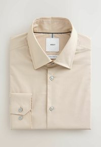 Chemise beige à manches longues, soigneusement pliée avec un poignet boutonné et un col pointu, étiquetée "NEXT" à l'intérieur du col.